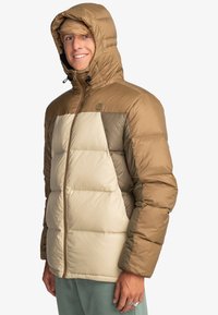 Pufferjack in bruin en crème, met een capuchon, doorgestikte design en een rits aan de voorkant. Gemaakt van lichtgewicht, waterafstotend materiaal.
