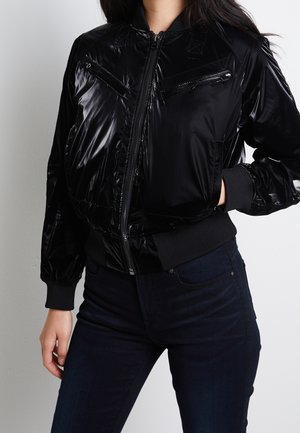 Femme portant une veste bomber noire brillante zippée avec des poches poitrine zippées et des poignets et ourlet côtelés, associée à un jean foncé.