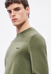 Zöld kötött pulóver bordázott textúrájú ujjakkal, kerek nyakkivágással, és fekete "BOSS" logóval a mellkason. Egységes, sima felület.