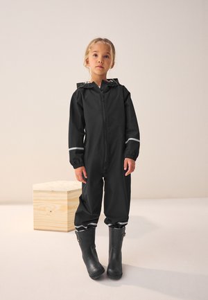 Schwarzer wasserdichter Overall mit elastischen Bündchen, Frontreißverschluss und reflektierenden Akzenten, kombiniert mit schwarzen Gummistiefeln. Einfarbiger Hintergrund.
