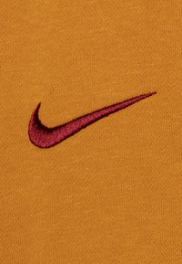 Nike-logo broderet i burgunder på et tekstureret, sennepsgult stof med et glat overflademønster uden yderligere udsmykninger.