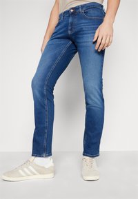 Jeans de denim azul con un corte ajustado, que presentan una textura desgastada, costuras en contraste, cinco bolsillos y un cierre de botón. Llevados con zapatillas ligeras.