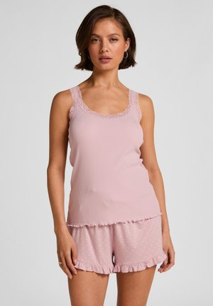 Vrouw draagt lichtroze hemdje met kanten afwerking en bijpassende polkadot gerimpelde korte broek, staand tegen een effen grijze achtergrond.