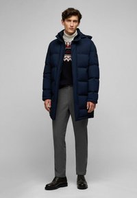Marineblå puffjakke med høj krave, med et quiltet design, båret over en mønstret trøje og grå bukser; sorte snørebåndssko fuldender outfittet.