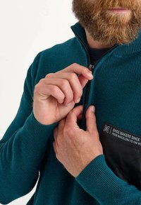 Sweat à zip couleur sarcelle avec un tricot texturé et un panneau en tissu noir portant un logo. Les mains du mannequin ajustent la fermeture éclair au niveau du col.