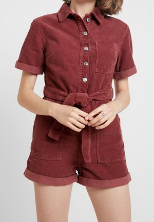 Femme portant une combishort en velours côtelé rouge à manches courtes, boutonnée à l'avant, avec une poche poitrine et une ceinture à nouer, les mains jointes à la taille.