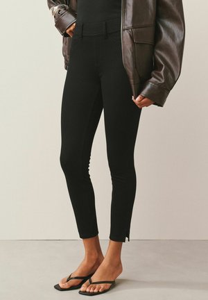 Frau trägt schwarze, eng anliegende Hose mit Knöchelschlitzen, schwarzes Tanktop, braune Lederjacke und schwarze Zehensandalen, steht vor neutralem Hintergrund.