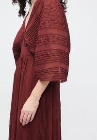 Burgunderfarget kjole med teksturert, plissért design. V-hals og vide, flytende ermer. Lett stoff med glatt finish.