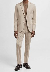 Abito elegante beige con blazer a due bottoni, taschino sul petto singolo e pantaloni con frontale piatto. Indossato sopra una maglietta marrone chiaro e mocassini marroni.