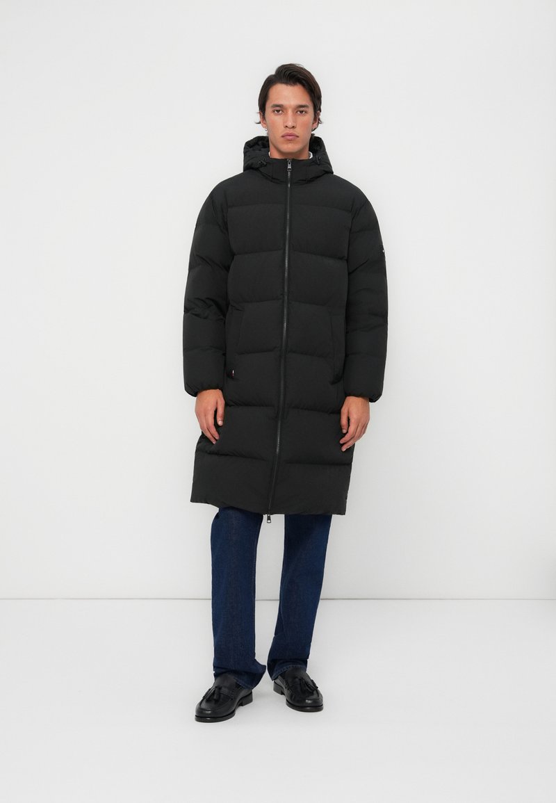 Zwarte pufferjas met horizontale quilt, rits aan de voorkant en capuchon. Vergezeld van donkerblauwe jeans en zwarte instappers op een witte achtergrond.