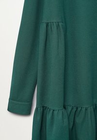 Robe à manches longues vert foncé avec des détails de coutures froncées à la taille et près des poignets sur un fond uni clair.