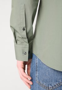 Cămașă verde deschis cu mâneci lungi și manșoane cu nasturi, combinată cu blugi din denim albastru, cu un aspect decolorat.