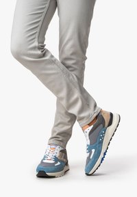 Blaue und graue Sportschuhe bestehen aus Mesh- und Wildledermaterialien, haben orangefarbene Akzente und eine gemusterte Zwischensohle. Kombiniert mit hellgrauen, schmal geschnittenen Hosen.
