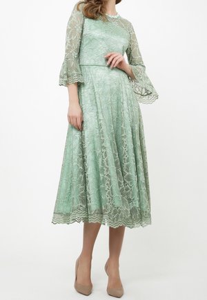 Woman wearing a mint green lace midi dress with bell sleeves and scalloped edges, paired with beige high-heeled shoes.