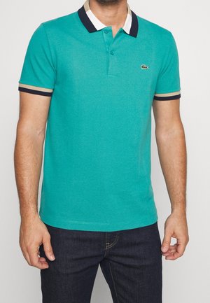 Homme portant un polo à manches courtes couleur sarcelle avec un col blanc et noir et un logo sur le côté gauche de la poitrine, assorti à un jean bleu foncé.