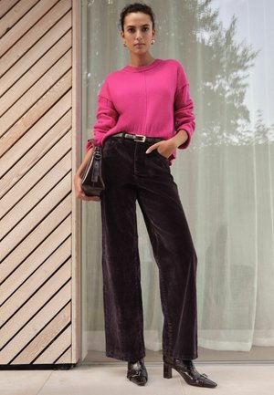 Fuchsia gestrickter Pullover mit fallenden Schultern, kombiniert mit dunklen Cordhosen in weiter Form. Dazu eine strukturierte braune Handtasche und schwarze High Heels.