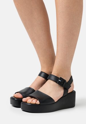 Mustat nahkaiset platform-sandaalit, joissa on avoin kärki, nilkkavyö ja solkiyksityiskohta; suunniteltu paksulla, teksturoidulla wedge-pohjalla.