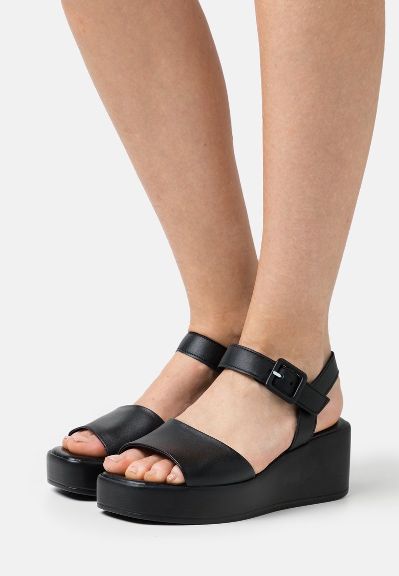 Mustat nahkaiset platform-sandaalit, joissa on avoin kärki, nilkkavyö ja solkiyksityiskohta; suunniteltu paksulla, teksturoidulla wedge-pohjalla.