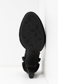 Semelle d'une chaussure à talon haut noire à bout pointu avec un motif floral, appuyée contre un mur d'angle blanc.