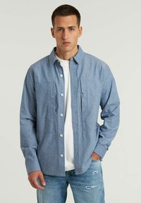 Camicia azzurra chiara con bottoni, realizzata in tessuto strutturato, con due tasche sul petto. Indossata sopra una t-shirt bianca dal taglio casual e con le maniche arrotolate.