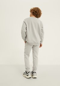Vaaleanharmaa sydänasu, joka koostuu crewneck-hupparista ja siihen sopivista joggereista. Materiaali on pehmeää ja hihansuissa on ribborakenteet. Mustat ja hopeiset lenkkarit.