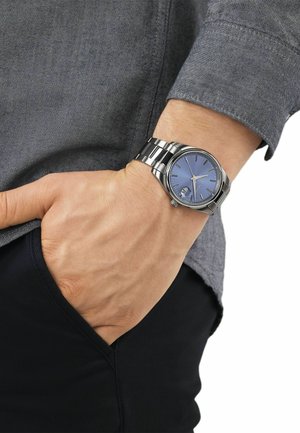 Left wrist with silver metal watch featuring a blue dial and date window, hand resting in black pants pocket, wearing gray long-sleeve shirt.