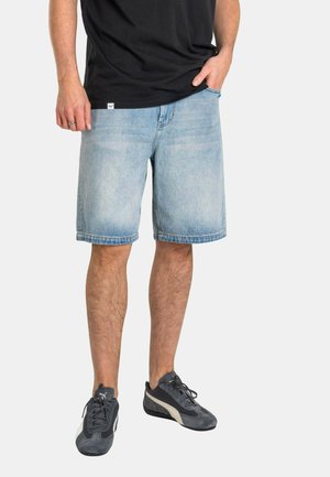 Mann trägt hellblaue Jeansshorts, schwarzes T-Shirt und dunkelgraue Puma-Sneaker, steht mit einer Hand in der Tasche.