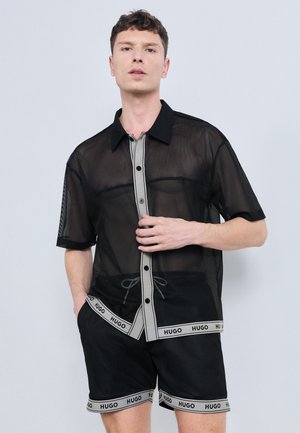 Tânăr purtând o cămașă neagră transparentă cu mâneci scurte și nasturi, împreună cu pantaloni scurți asortați cu margini gri, etichetată „HUGO” pe un fundal gri.