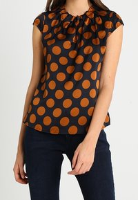 Kvinna i kortärmad svart blus med stora oranga prickar, tillsammans med mörkblå jeans, som står mot en enfärgad bakgrund.