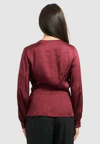 Blusa di satin bordeaux con maniche lunghe, scollatura rotonda e vestibilità aderente in vita, abbinata a pantaloni neri. Texture liscia.