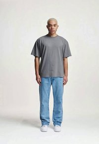 T-shirt oversized cinza com mangas curtas, combinada com jeans de tom azul claro. Modelo a usar ténis brancos, de pé contra um fundo claro.