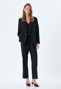 Schwarzer maßgeschneiderter Anzug mit einem strukturierten Blazer mit Revers, einer taillierten Weste mit Knöpfen und geraden Hosen. Glänzende schwarze Absatzschuhe.
