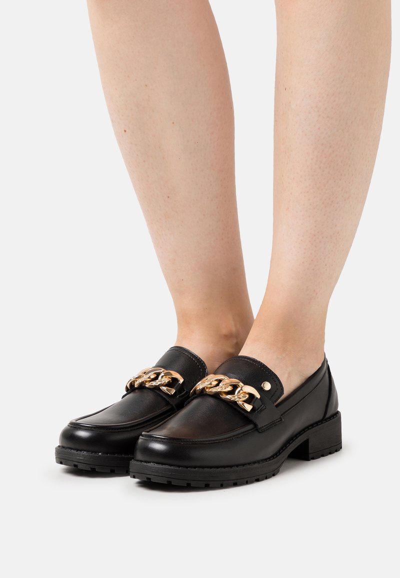 Mocassins en cuir noir avec un gros détail de chaîne dorée portés pieds nus sur un fond blanc.