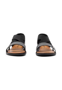 Czarne sandały z krzyżującymi się paskami z elastycznego materiału, otwartym palcem, gładką brązową wkładką i płaską czarną podeszwą.