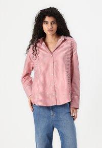Camisa às riscas vermelha e branca, com colarinho, mangas longas e um corte descontraído, combinada com jeans azuis.