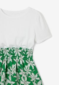 Vitt kortärmat bomullstopp med en smockad grön blommig kjol som har vita blommor och gröna prickiga detaljer.