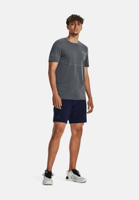 Camisa de deporte gris de manga corta, cuello redondo y pequeño logo azul. Lleva puestos unos pantalones cortos de color azul marino oscuros y zapatillas deportivas blancas.