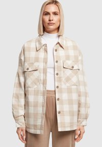 Veste-chemise à carreaux beige et blanc avec une fermeture boutonnée sur le devant, deux poches poitrine et des manches longues. Texture douce, coupe décontractée et col arrondi.