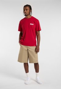 Camiseta roja de manga corta con logo blanco, combinada con pantalones cortos beige hasta la rodilla. El modelo lleva zapatillas blancas y calcetines blancos contrastantes.