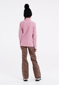 Giacca in pile rosa chiaro con colletto alto, abbinata a pantaloni marroni a zampa d'elefante con un motivo a stampa leopardata nera. Berretto lavorato a maglia nero con pom-pom.