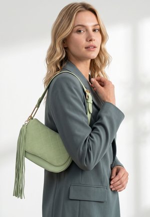 Donna con capelli biondi ondulati che indossa un blazer verde smorzato, porta una borsa a tracolla trapuntata verde chiaro con dettagli di lunghe nappine.