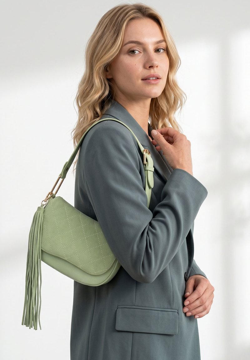 Femme aux cheveux blonds ondulés portant un blazer vert atténué, portant un sac à bandoulière matelassé vert clair avec des détails de longs pompons.