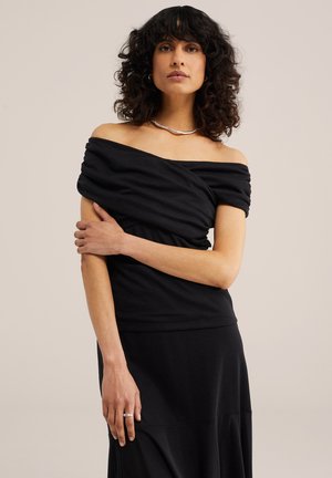 Kobieta o kręconych włosach, ubrana w czarny top odsłaniający ramiona i czarną spódnicę, ozdobiona srebrnym naszyjnikiem i pierścionkiem, stojąca z jedną ręką skrzyżowaną na piersi.