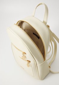 Beige faux skinnsekk med gullglider, med en frontlomme og justerbare stropper. Innvendig er den foret med stoff.