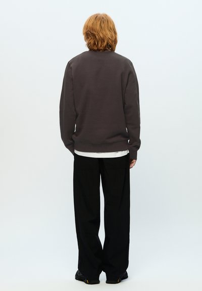 Dark gray sweatshirt with a relaxed fit, ribbed cuffs, and hem, worn over a white shirt. Paired with loose black trousers and black footwear.