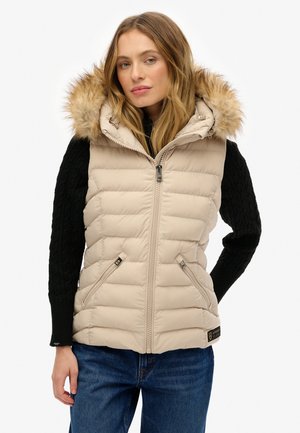 Gilet en duvet beige avec capuche en fourrure amovible, fermeture éclair sur le devant, texture matelassée et deux poches latérales, porté sur un pull noir.
