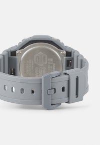 Gray silicone watch strap with a stainless steel back, featuring a pin buckle and adjustment holes for sizing. Textured surface for grip.