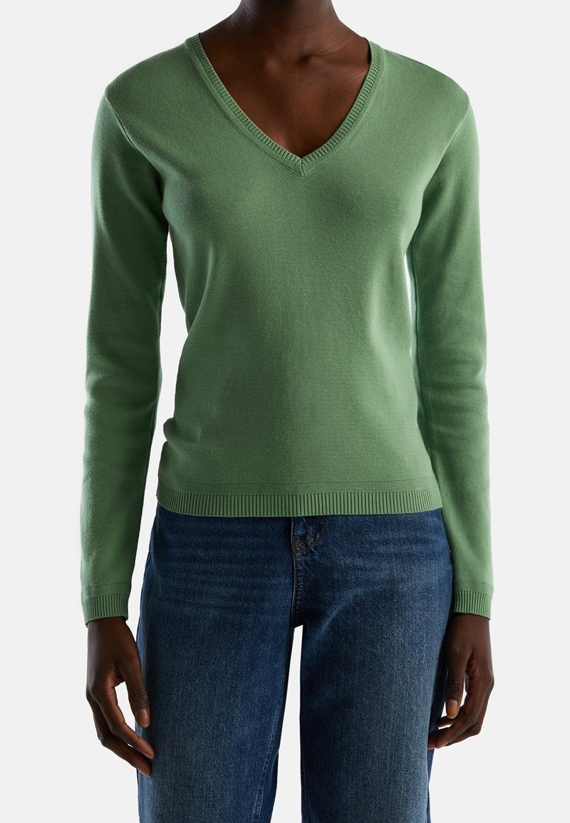 Grön långärmad tröja med V-ringning, ribbad halsringning och fåll, kombinerad med blå denimjeans, som visar en slät stickad struktur.