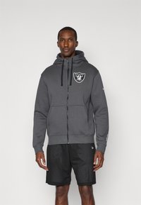 Szary hoodie na zamek wykonany z miękkiego materiału, z logo z przodu, regulowanymi sznurkami oraz kieszeniami typu kangur. Noszony z czarnymi szortami z siateczki.