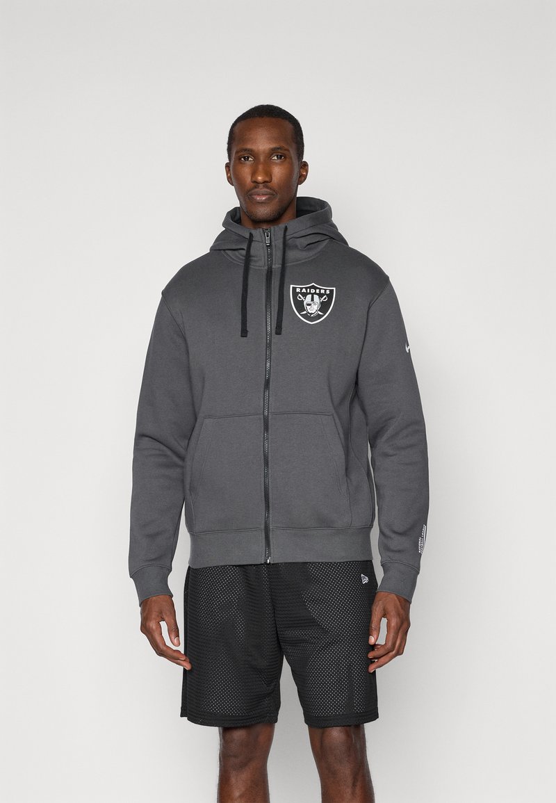 Szary hoodie na zamek wykonany z miękkiego materiału, z logo z przodu, regulowanymi sznurkami oraz kieszeniami typu kangur. Noszony z czarnymi szortami z siateczki.